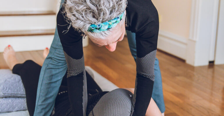 A person helping another with a floor stretching exercise indoors.