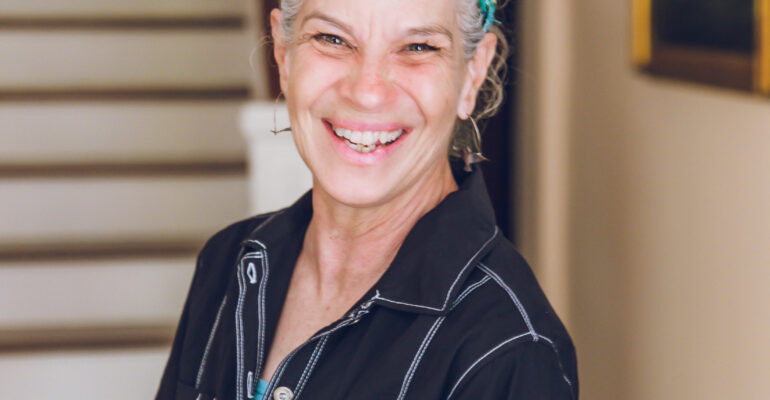 Smiling woman with a blue headband stands indoors near stairs.